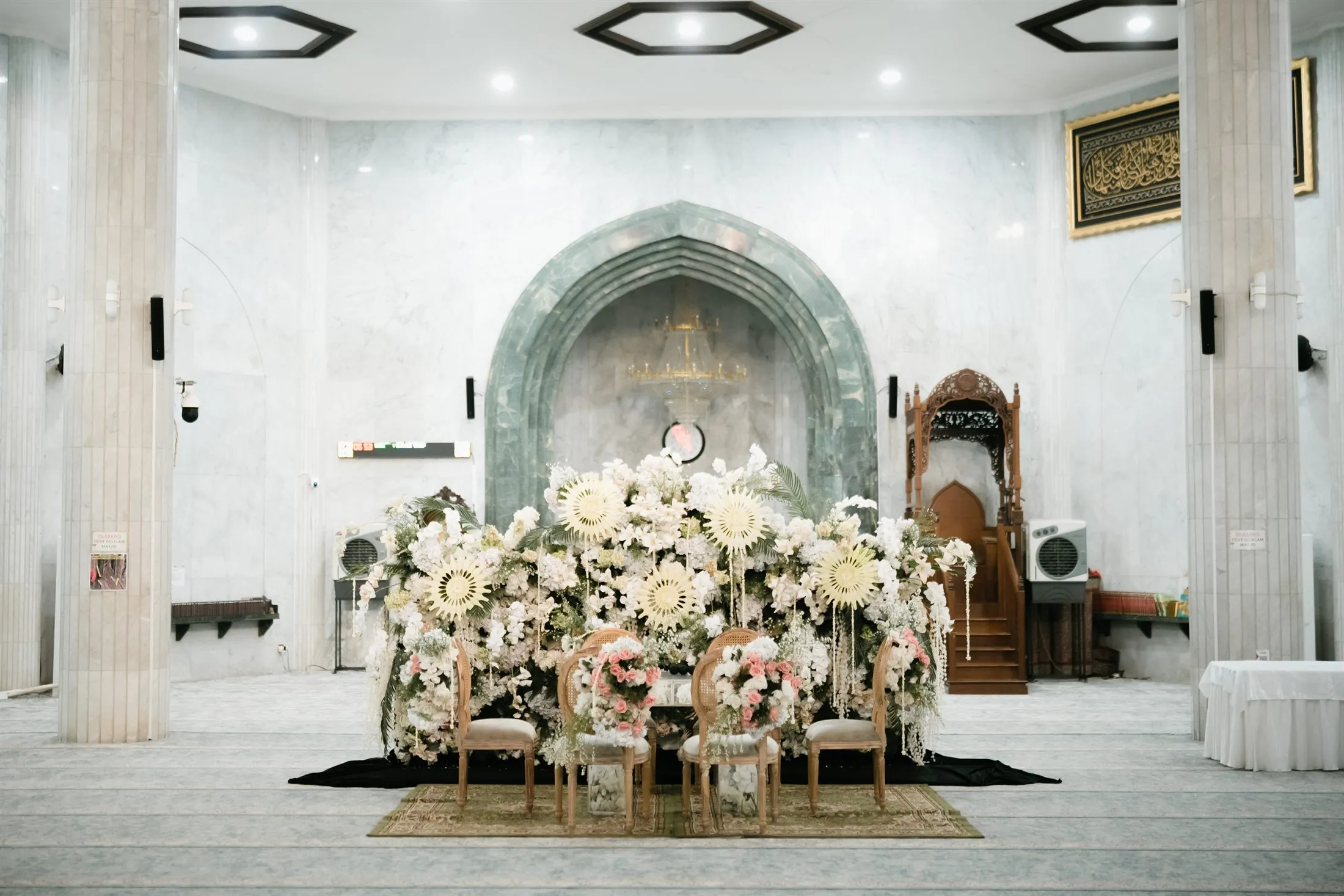 Dekorasi akad nikah dengan latar mihrab dan rangkaian bunga putih di Masjid At-Taqwa Sriwijaya