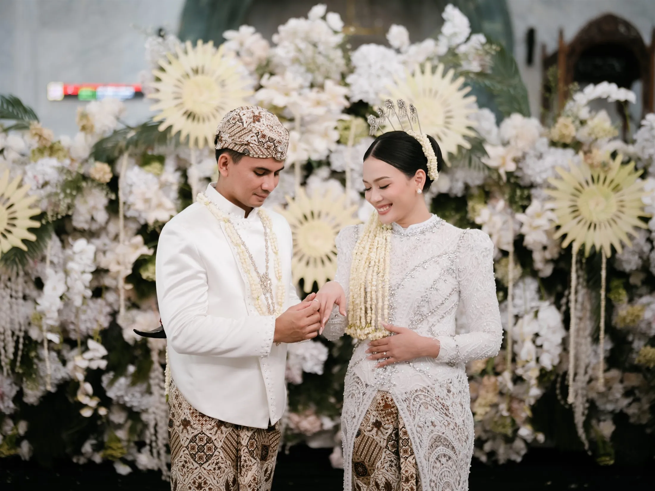 Pasangan pengantin Astiara dan Idul mengenakan busana adat nuansa putih gading dengan latar bunga